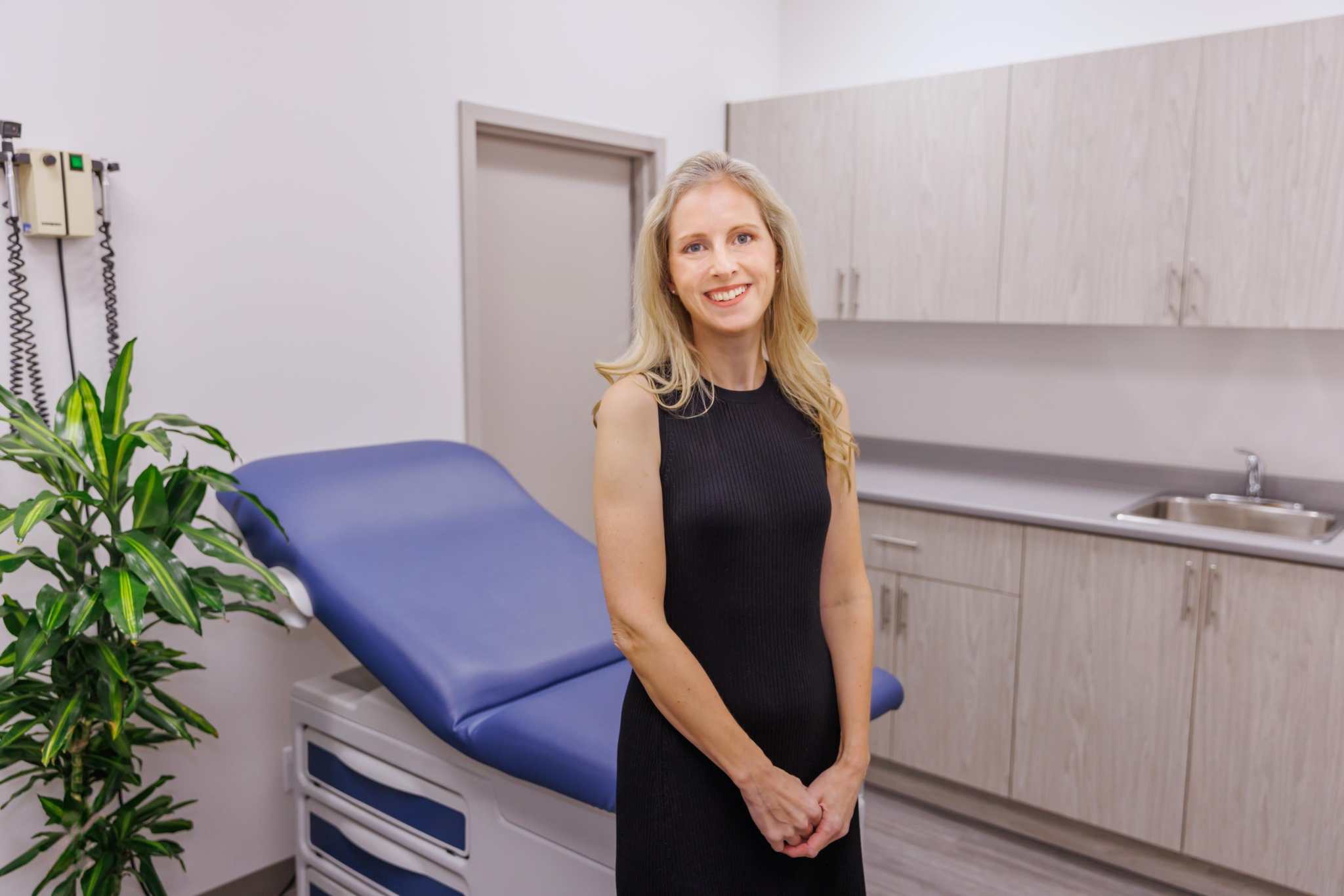 Woman standing in modern medical aesthetics clinic room with blue treatment chair and light wood cabinets at Merrion Medical Aesthetics in Oakville.