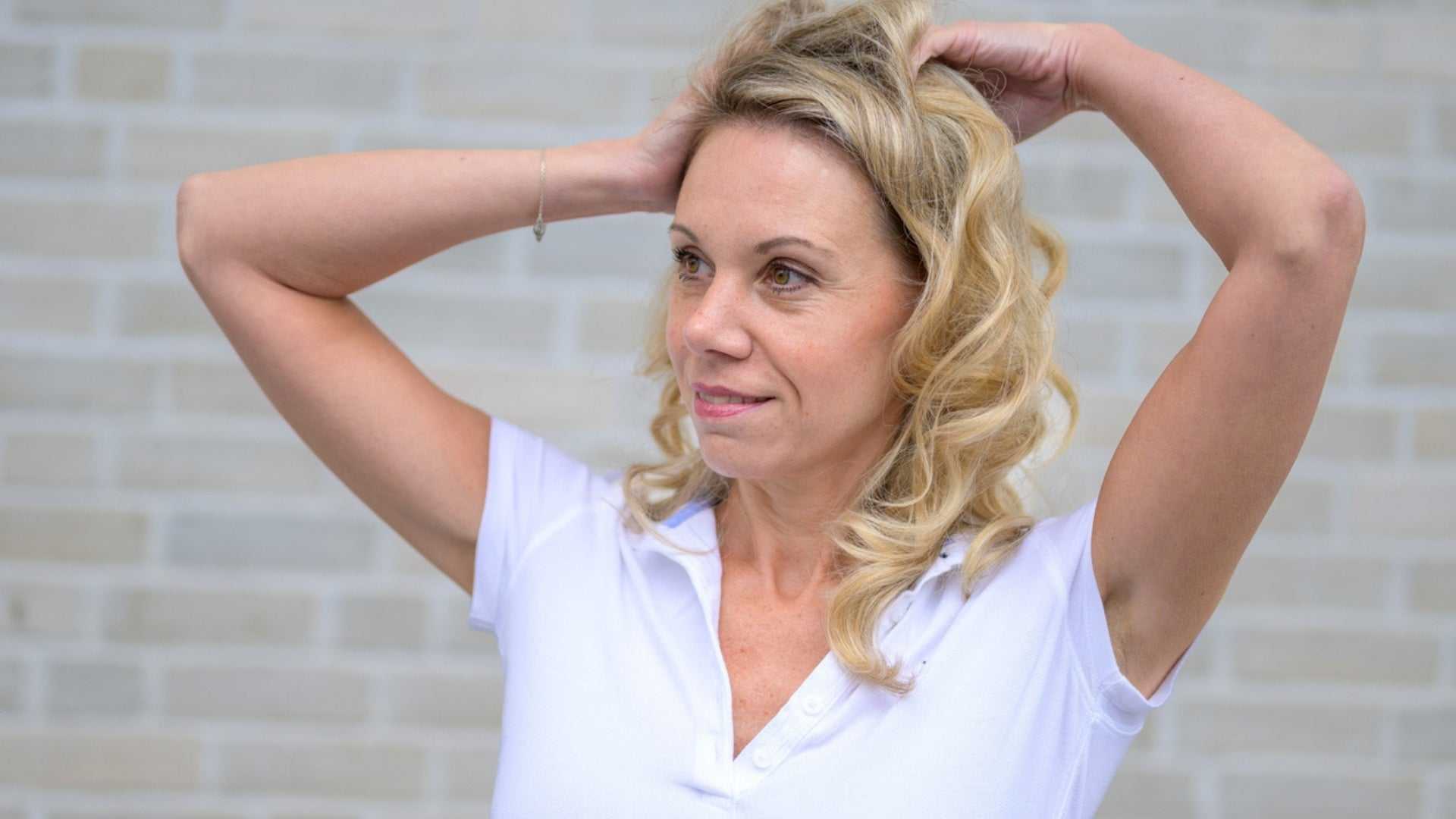 middle-aged woman with blonde curly hair wearing a white shirt posing against a light brick wall for Botox treatments in Burlington advertisement