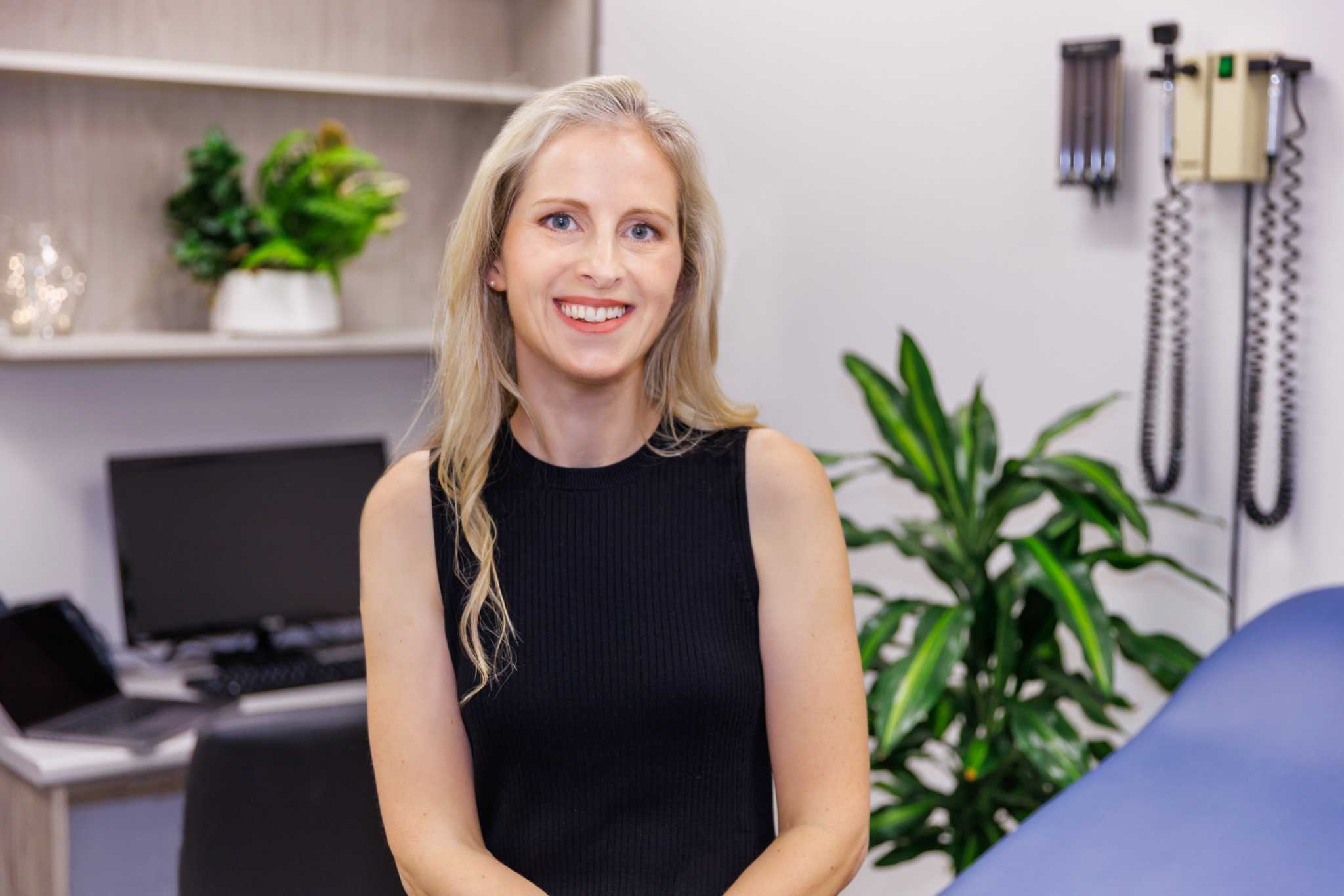 Female medical professional smiling in clinic room with plants and medical equipment in background