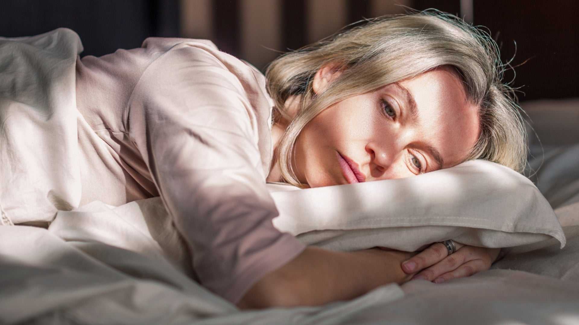 Woman with gray hair lying on bed looking thoughtful, representing menopause and its effects on skin health and well-being