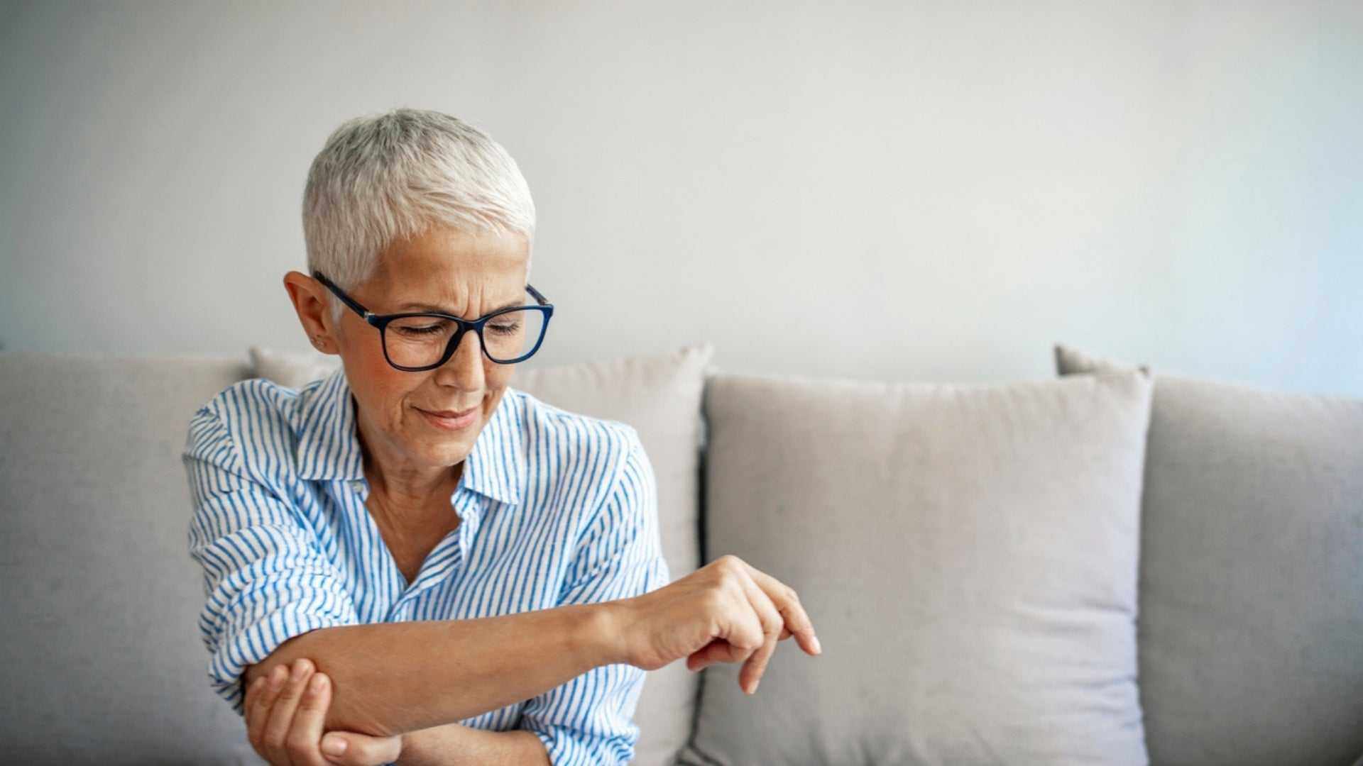 Mature woman with short gray hair and glasses touching her elbow in discomfort on a couch, highlighting bone health concerns in perimenopause.