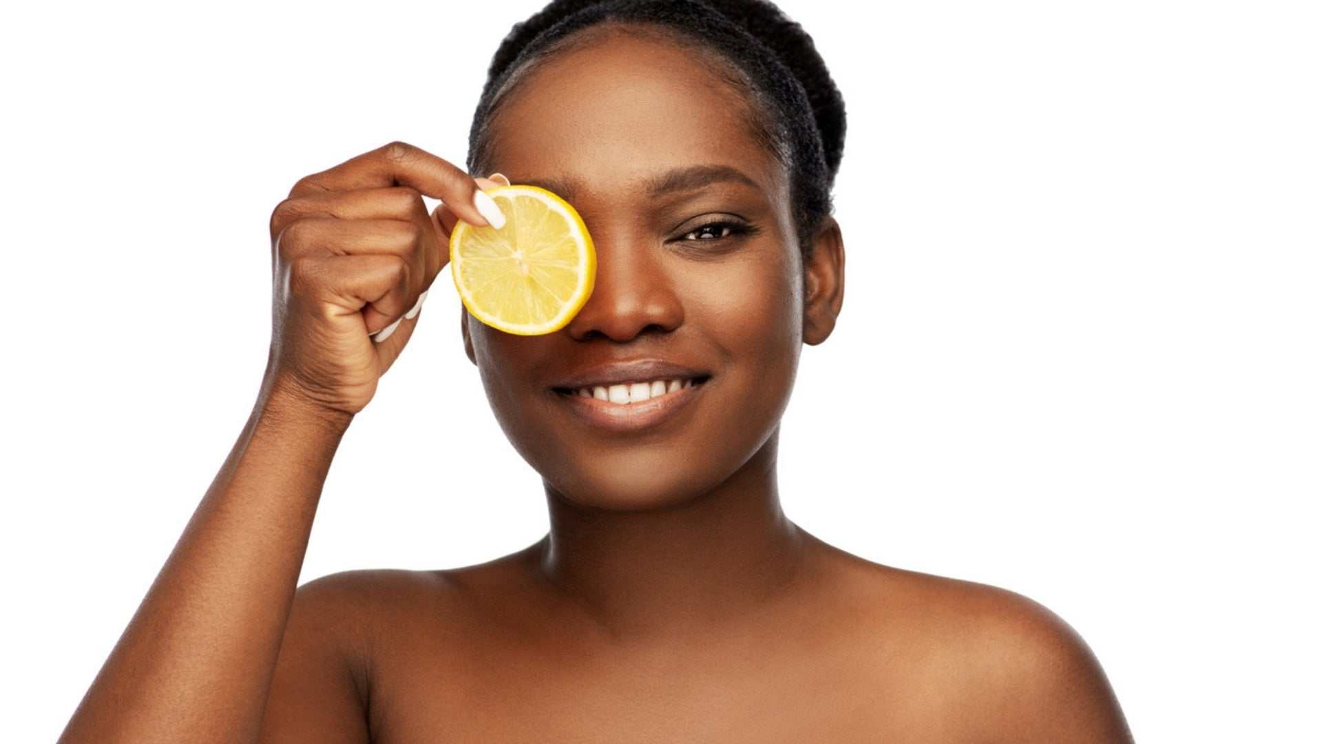 Smiling woman holding lemon slice over eye representing vitamin C skincare benefits for radiant healthy skin