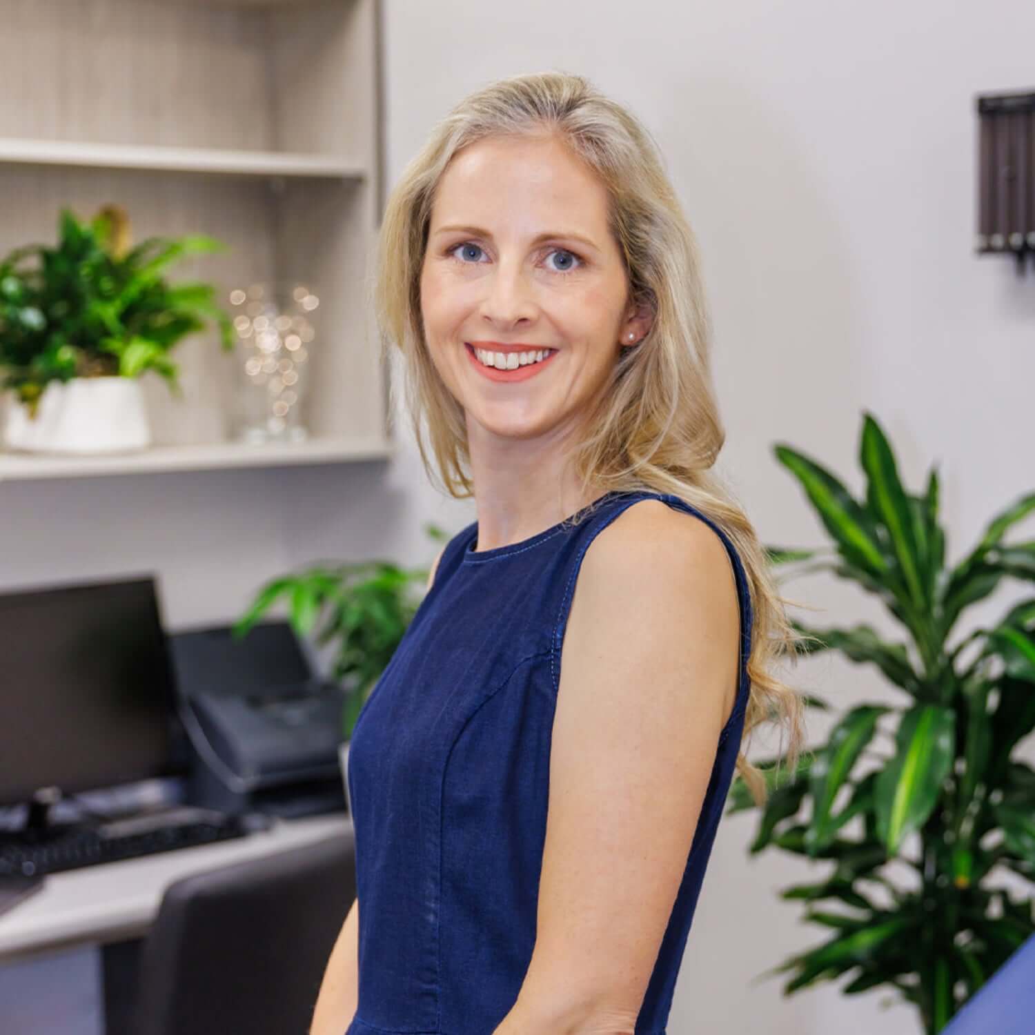 Woman in a blue dress standing in an office setting with plants and office equipment.