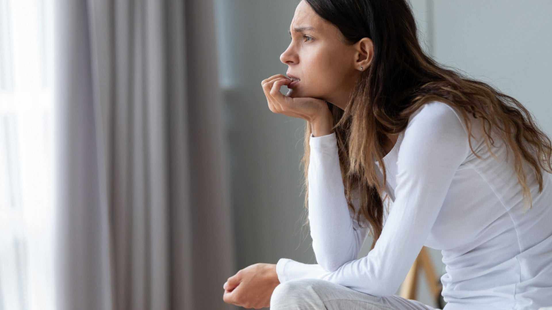 How Stress Affects Skin: stressed woman sitting by window, worried expression, highlighting cortisol-related acne and rosacea flare-ups