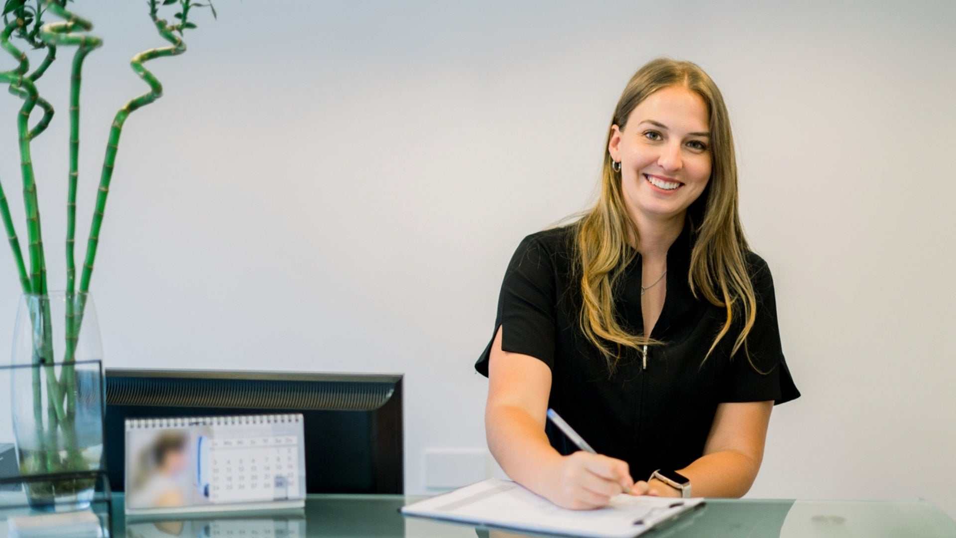 Smiling woman in black shirt writing at a reception desk in a medical or cosmetic clinic environment