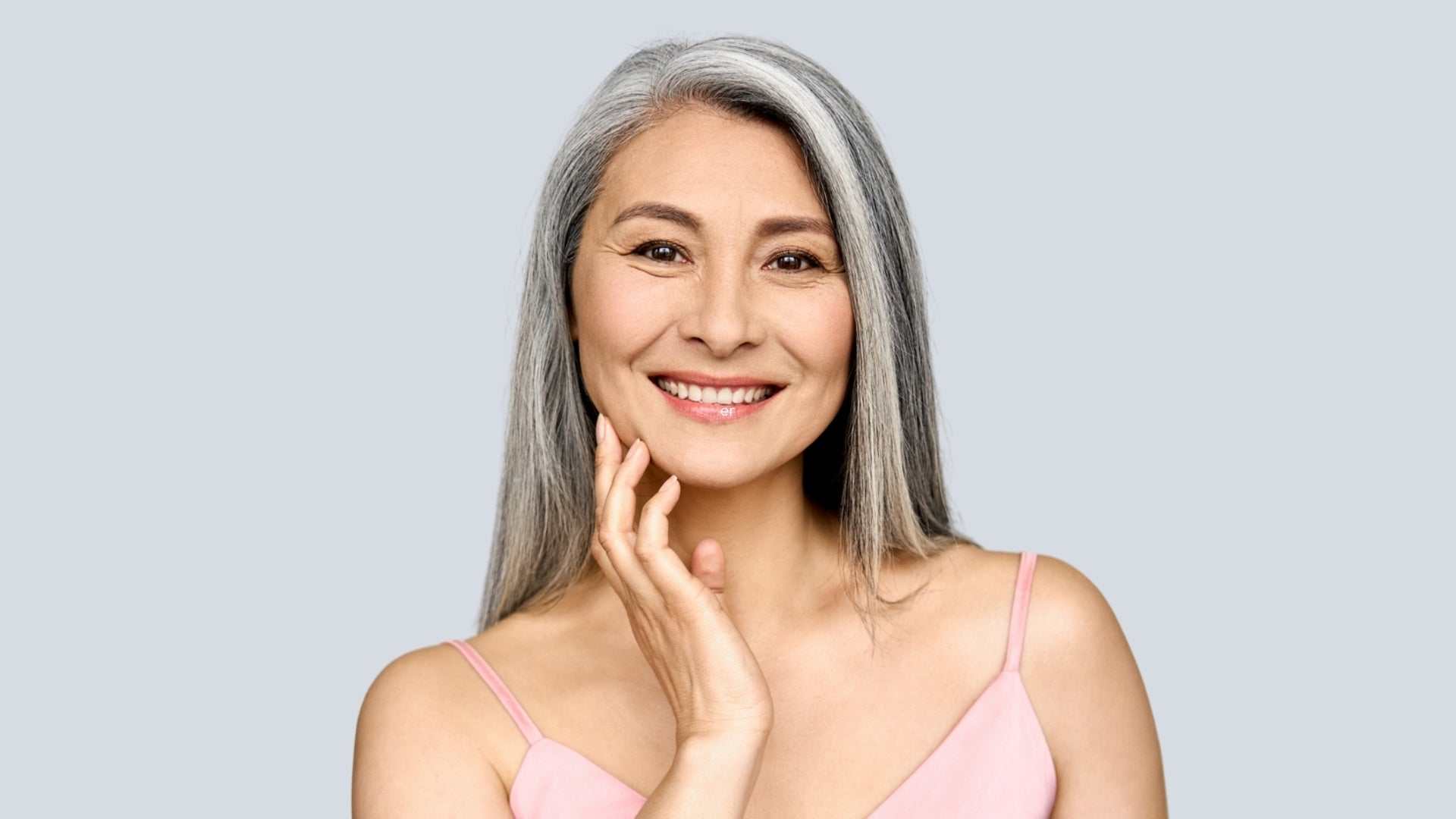 Smiling middle-aged woman with long gray hair touching her face, representing skin tightening treatments for aging and loose skin.