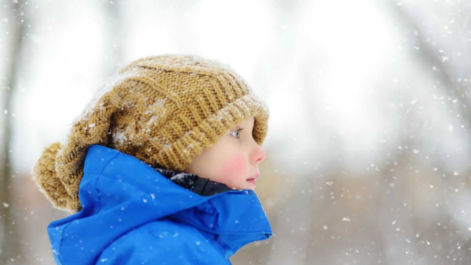 Young child with rosy cheeks wearing a blue winter jacket and knitted hat in falling snow, illustrating winter skincare routine protection