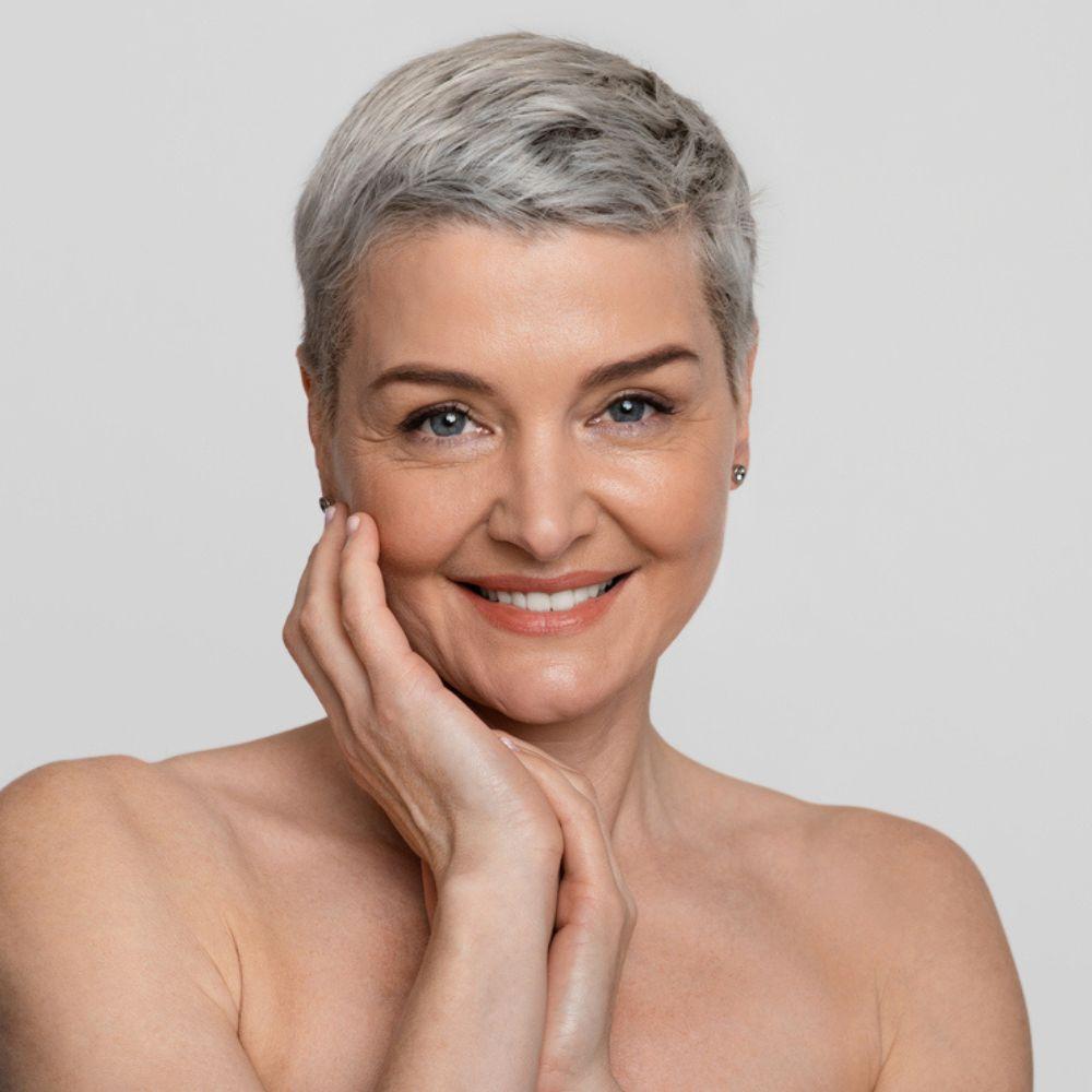 Woman with short gray hair smiling against a plain background. Mature woman after skin rejuvenation treatment showing improved skin texture and firmness - comprehensive anti-aging laser program at Merrion Medical Aesthetics Oakville
